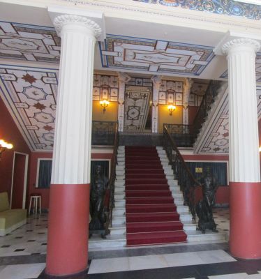 A photo from a lower perspective looking up the Achilleion's main staircase, with a grand chandelier visible in the distance, creating a sense of following in Empress Sisi's footsteps.