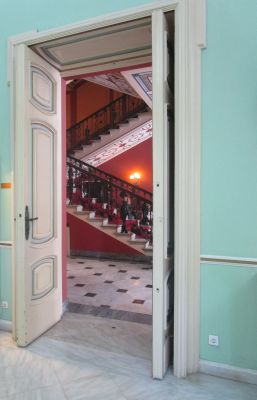 A doorway framed by ornately decorated walls offers a view of the Achilleion Palace's grand staircase.